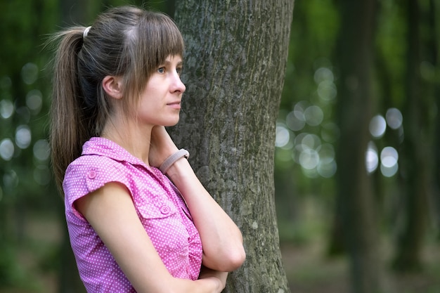 Young woman leaning to tree trunk in summer forest.