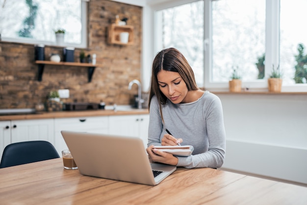 Young woman learning online with laptop and taking notes in a notebook at home.