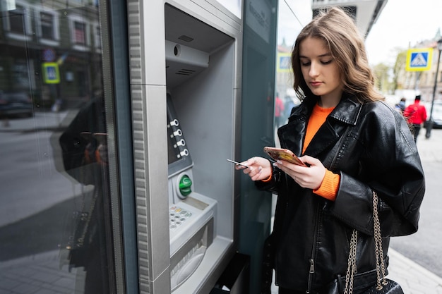 Young woman in leather jacket inserting a credit card to ATM outdoor while looking on smartphone