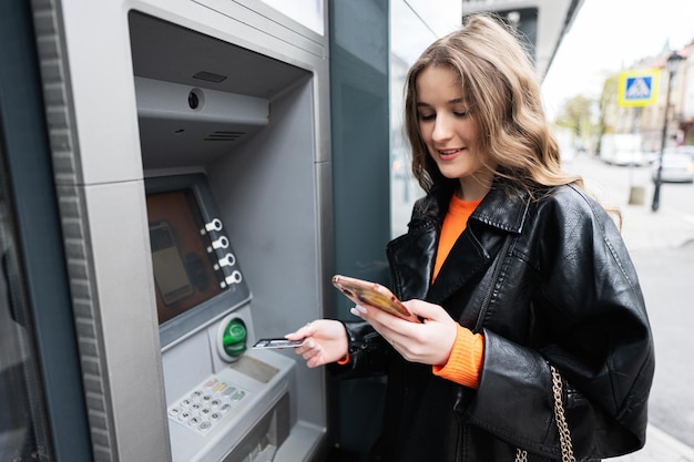 Young woman in leather jacket inserting a credit card to ATM outdoor while looking on smartphone