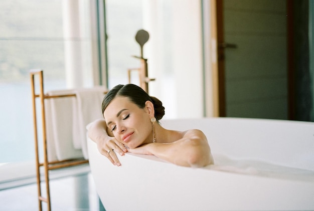 Young woman lies in a bathtub with foam with her head in her hands