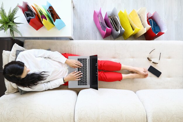 Young woman lies on couch with laptops on her lap with many paper bags on floor in apartment