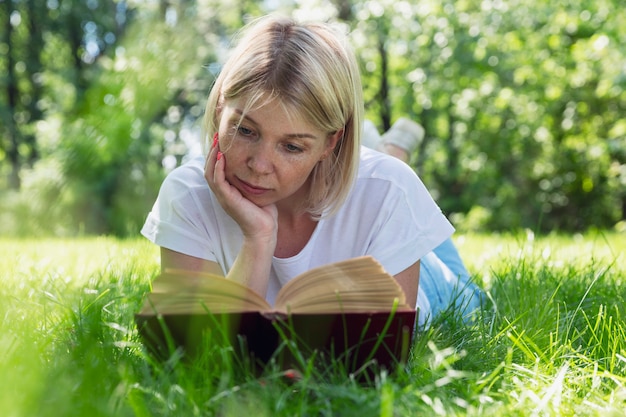 젊은 여자가 공원의 잔디에 누워 책을 읽습니다. 주근깨가 있는 예쁜 금발. 화창한 여름날에 휴식을 취하십시오.