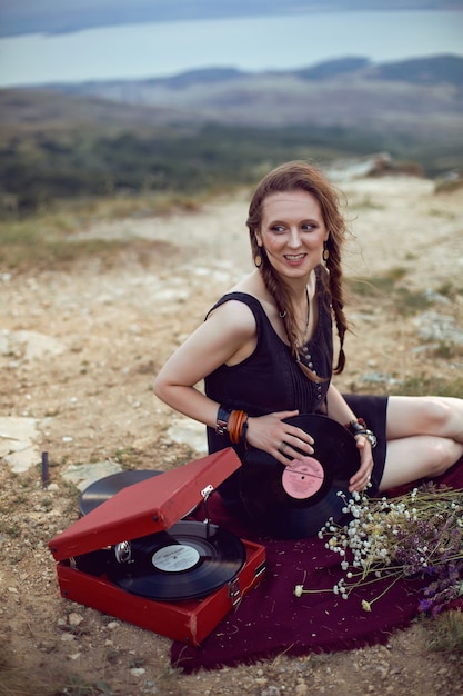 Young woman lies in nature in a black dress next to an old gramophone and listens to music