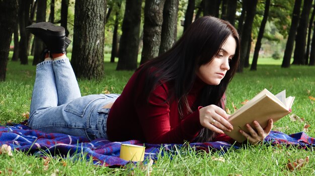 A young woman lies and reads her favorite book on a woolen plaid blanket or blanket in a city park on green grass on a pleasant sunny day. The concept of recreation and leisure, education and study.