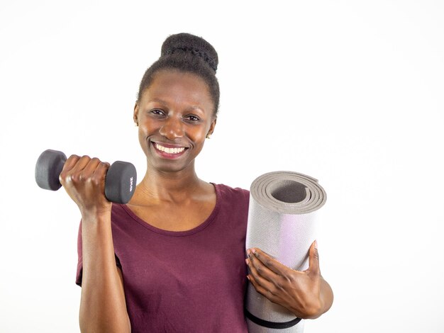 Young woman lifting a dumbbell while holds a yoga mat isolated on white background