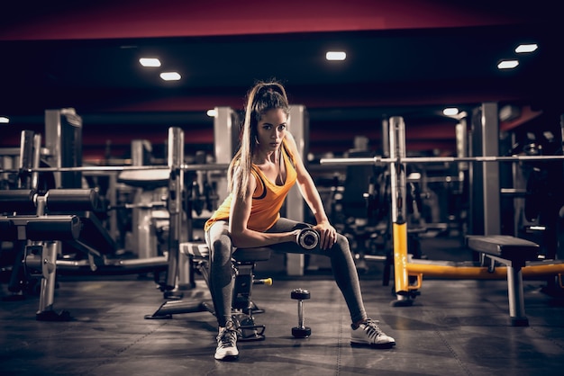 Young woman lifting weights and sitting on bench while looking at camera. Side light, gym interior.