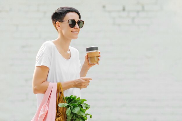 Young woman in light summer clothes with a eco bag of vegetables, greens and reusable coffee mug. Sustainable lifestyle. Eco friendly concept.