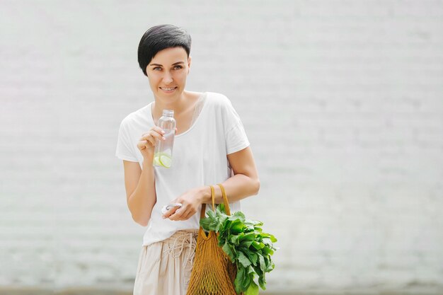 Young woman in light summer clothes with a eco bag of vegetables, greens and reusable water bottle.  Sustainable lifestyle. Eco friendly concept.