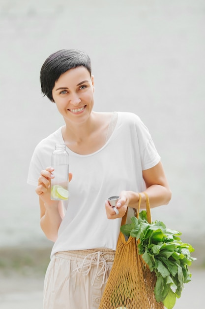 Молодая женщина в легкой летней одежде с эко-сумкой овощей, зелени и многоразовой бутылкой с водой. Устойчивый образ жизни. Экологичная концепция.