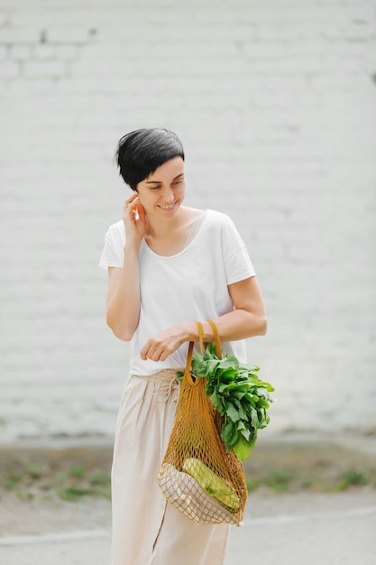 野菜と緑のエコバッグと明るい夏の服を着た若い女性。持続可能なライフスタイル。環境にやさしいコンセプト。