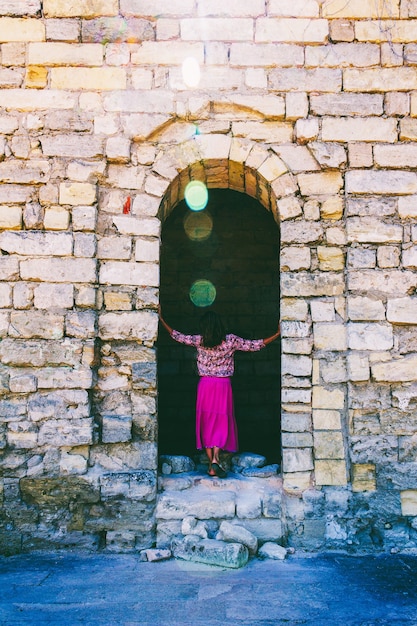 ライラックのスカートをはいた若い女性が、古代の要塞の廃墟に背を向けて立っています。