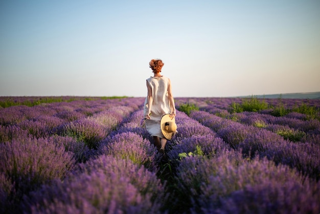 Young woman in linen dress holding straw hat in her hands running on the lavender field Back view