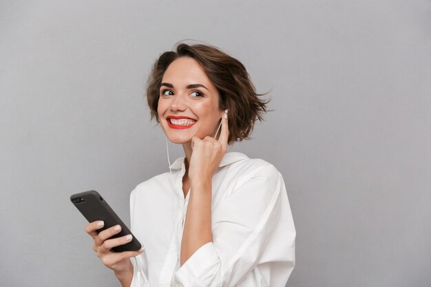 young woman listening to music on smartphone via earphones, isolated over gray wall