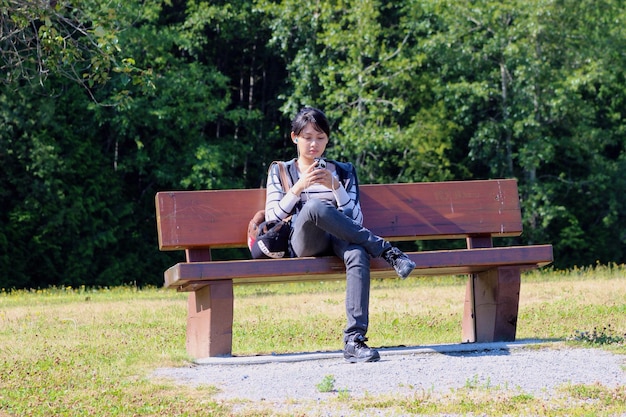 Young woman listening music through smart phone while sitting on bench against trees at park