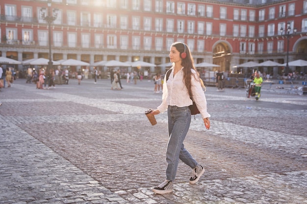 Young woman listening to music and walking in city with hot drink in hand