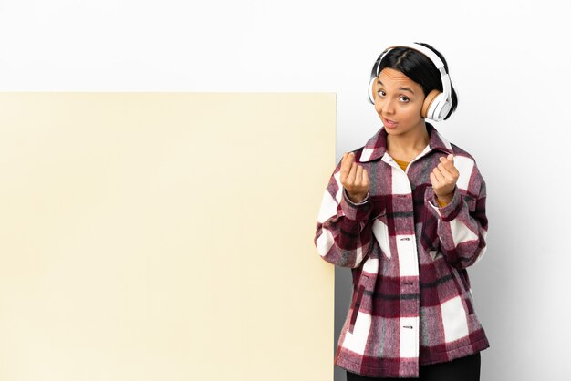 Young woman listening music with a big empty placard over isolated background making money gesture