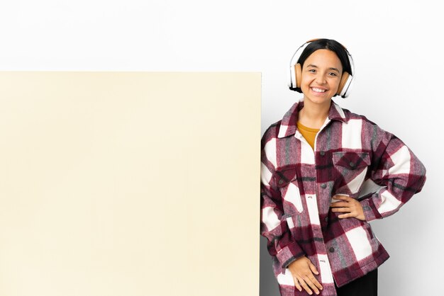 Young woman listening music with a big empty placard over isolated background posing with arms at hip and smiling