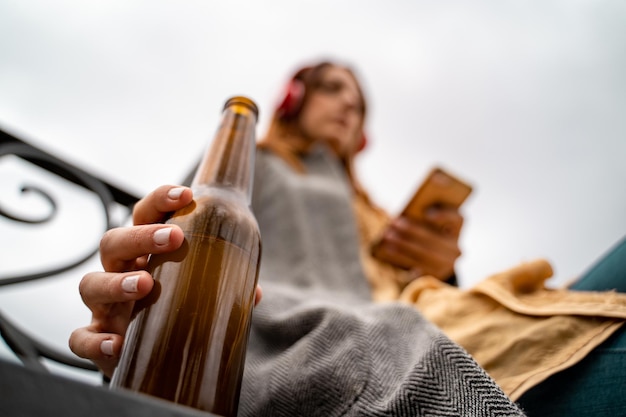 Foto giovane donna che ascolta la musica con le cuffie e lo smartphone bevendo birra nel parco focus sulla bottiglia di birra marrone con la mano su di essa volto sfocato