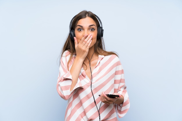 Young woman listening music with a mobile over isolated blue wall with surprise facial expression