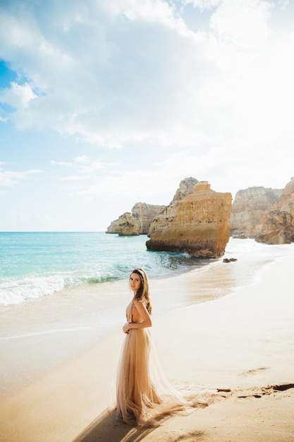 young woman in a long dress walking on beach near ocean.