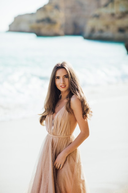 young woman in a long dress walking on beach near ocean.