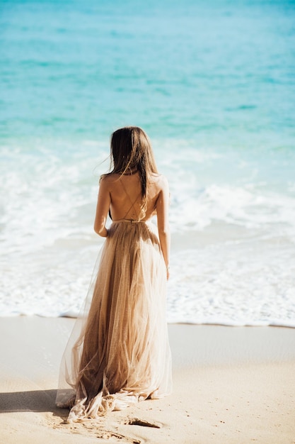 young woman in a long dress walking on beach near ocean.