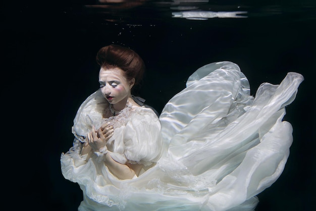 young woman in long white luxury dress underwater on the black background. Motion, art, winter