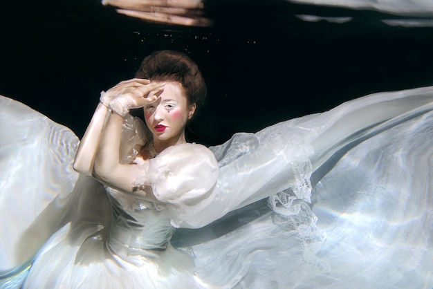 young woman in long white luxury dress underwater on the black background. Motion, art, winter