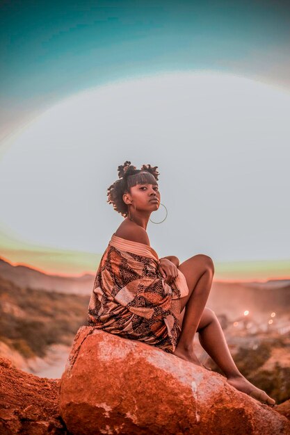 Young woman looking away while sitting on rock against sky