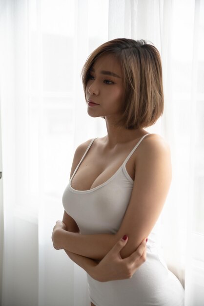 写真 家の窓のそばに立っている若い女性が目をそらしている