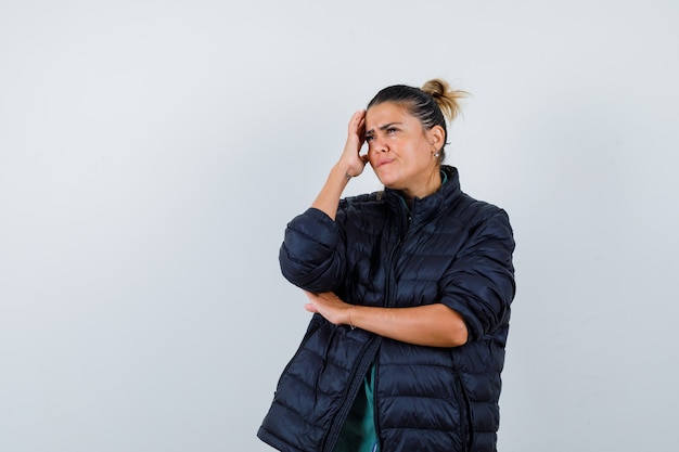 Young woman looking away with hand on head in puffer jacket and looking thoughtful. front view.