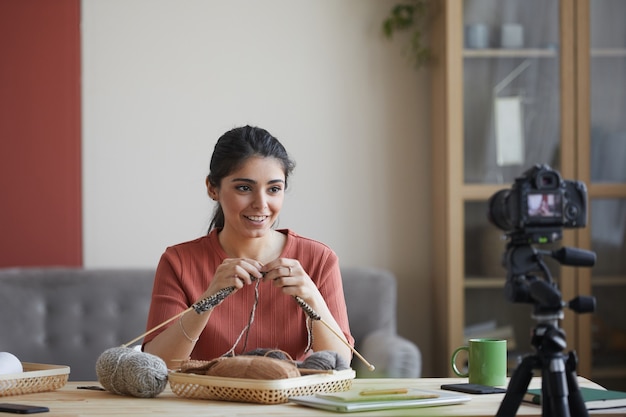 young woman looking at camera and teaching to knitting her followers during live broadcast