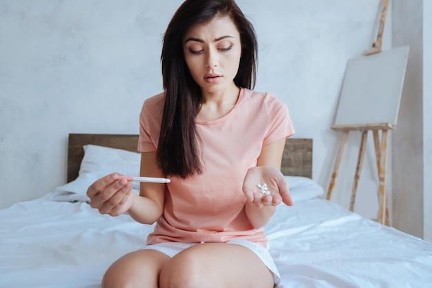 Young woman looking confused while checking a pregnancy test results and holding pills in her hand