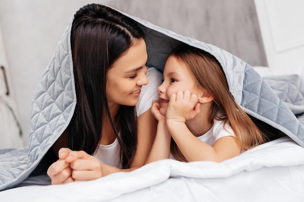 Young woman looking at her daughter while lying closely under blanket and feeling excited