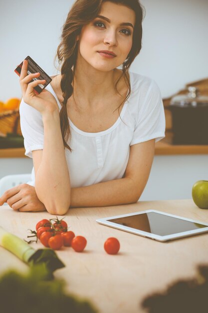 Молодая женщина ищет новый рецепт приготовления пищи на кухне. Домохозяйка делает покупки в Интернете с помощью планшетного компьютера и кредитной карты.