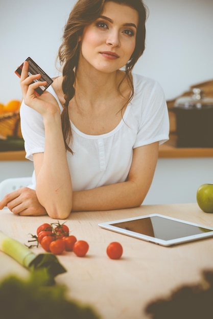 キッチンで料理するための新しいレシピを探している若い女性。主婦はタブレットコンピューターとクレジットカードでオンラインショッピングをしています。