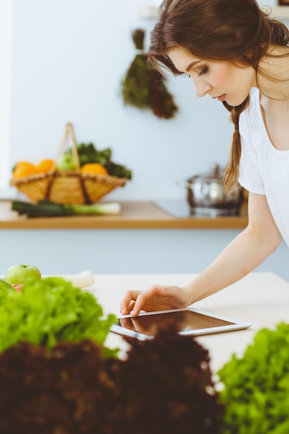 Молодая женщина ищет новый рецепт приготовления пищи на кухне. Домохозяйка делает покупки в Интернете с помощью планшетного компьютера и кредитной карты.