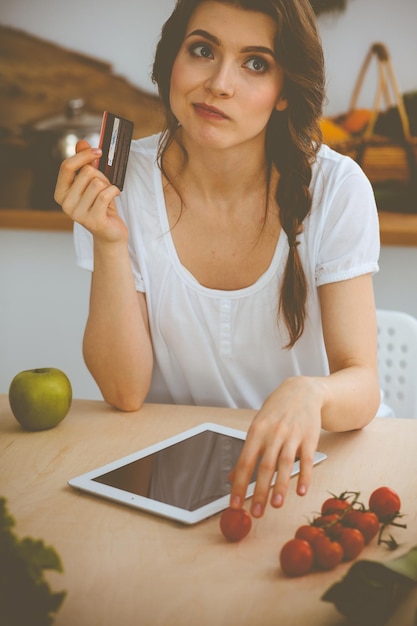 料理の新レシピを探している若い女性家庭主婦がタブレットコンピュータとクレジットカードでオンラインショッピングをしています