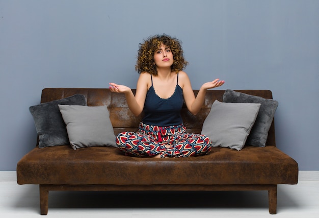 Foto giovane donna che sembra perplessa, confusa e stressata, chiedendosi tra diverse opzioni, sentendosi incerta seduta su un divano.