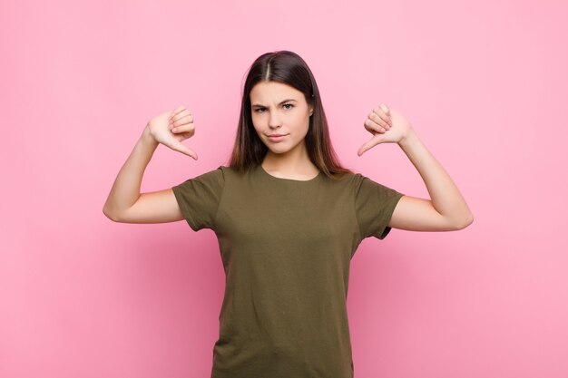 Young woman looking sad, disappointed or angry, showing thumbs down in disagreement, feeling frustrated on pink wall