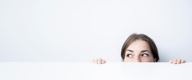 Giovane donna che guarda al lato che guarda fuori da sotto un tavolo bianco contro un muro bianco banner