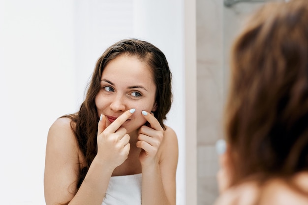 Giovane donna alla ricerca e spremere l'acne su una faccia davanti allo specchio. brutta ragazza con la pelle problematica, ragazza adolescente con i brufoli. cura della pelle. bellezza