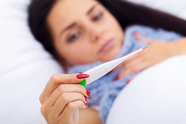 Giovane donna guardando il termometro mentre tenendolo in mano e sdraiato nel letto