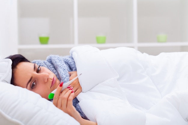 Young woman looking at thermometer while holding it in hands and lying in bed 