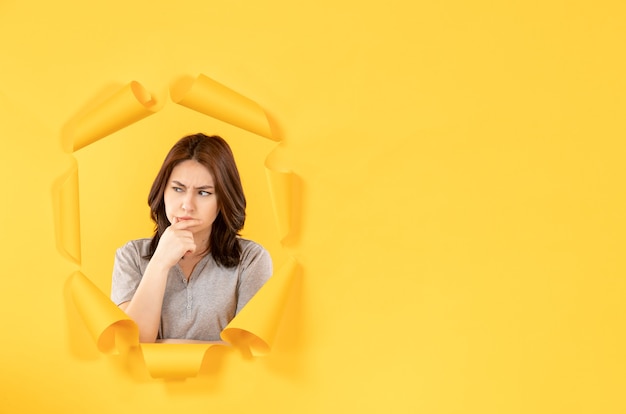 young woman looking through a hole in the yellow paper background window advertising spy