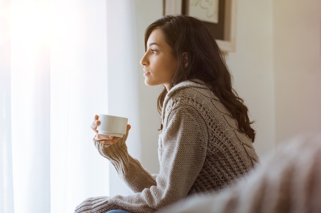 コーヒーを保持している窓ガラスを見ている若い女性