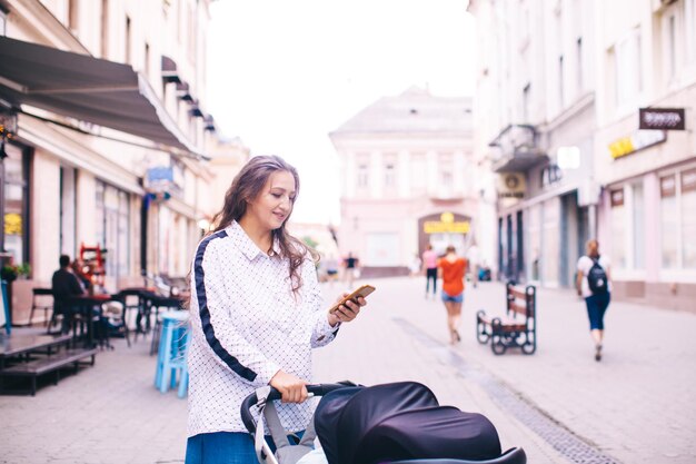 Photo young woman looks into her smartphone and walks with a baby stroller in the city center