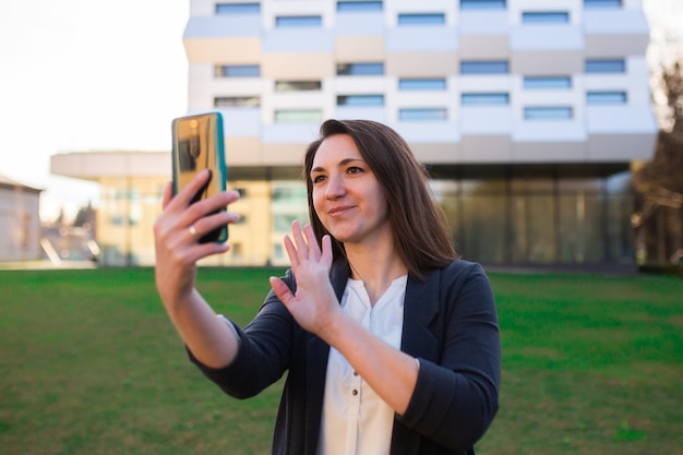 Young woman looks at the phone, talks on the phone, talks on video, smiles, waves, greets, on the street in the park, near the business center