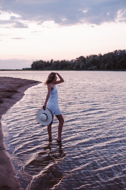 Una giovane donna guarda il tramonto una bella bionda snella felice è in piedi sulla riva del fiume in ...